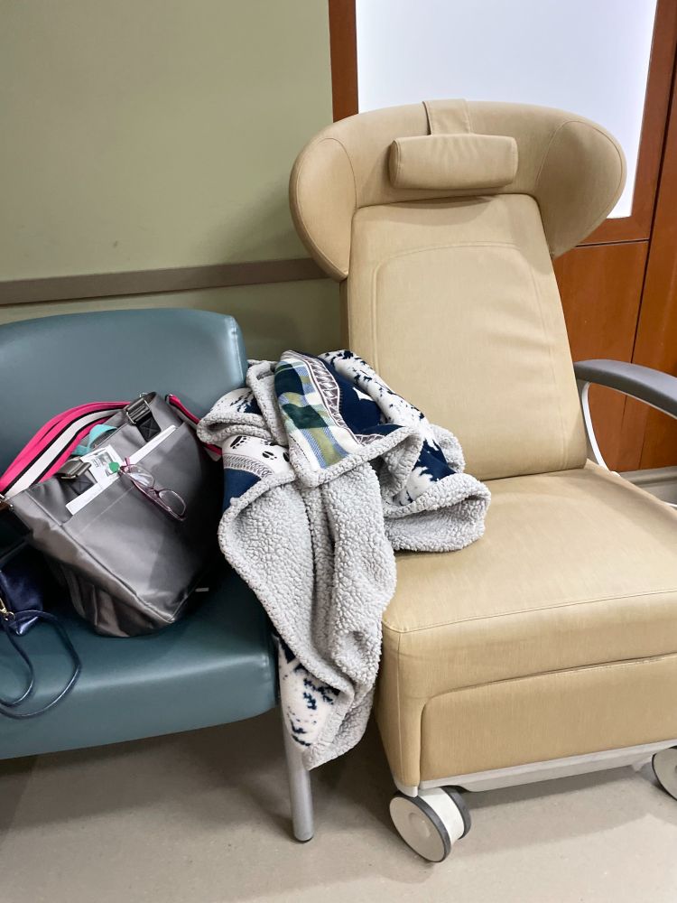 Chairs in a hospital room, one with laptop bag and purse. A blanket drapes between them over the chair arms.