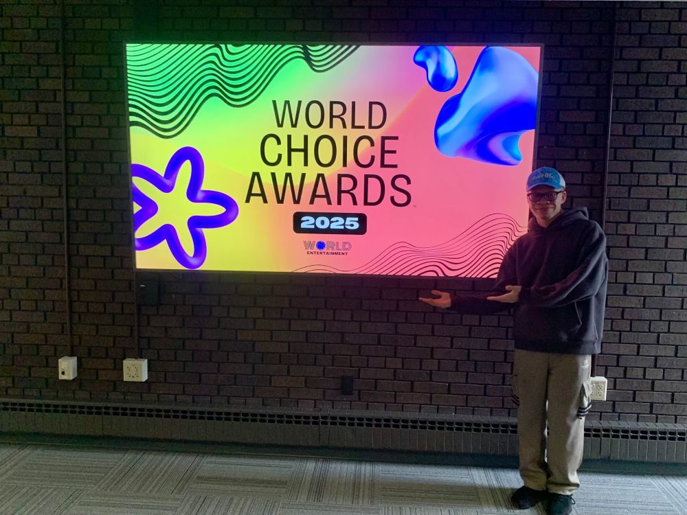 A picture of Paul Victor the creator of the World Choice Awards™ standing next to the 2025 Logo on a flat screen television hung up on the wall.