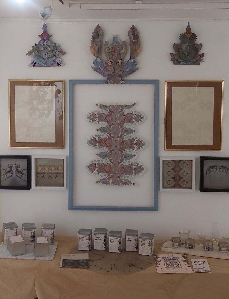 a large picture of a repetition pattern made from jelly fish is in the centre, on either side in a gold frame is a plant based ink screen print of a repetitive pattern from the reflection on the surface of water. above are three mirrored reflections of an individual story, then underneath the golden frames are 4 repeating patterns, from jelly fish showing a spirit and the basis of the large central painting, and patterns from seaweed that look like male and female abstract portraits. There is the top of a table with 14 teas laid out on reclaimed marble and recycled plastic, in large and small tins. 