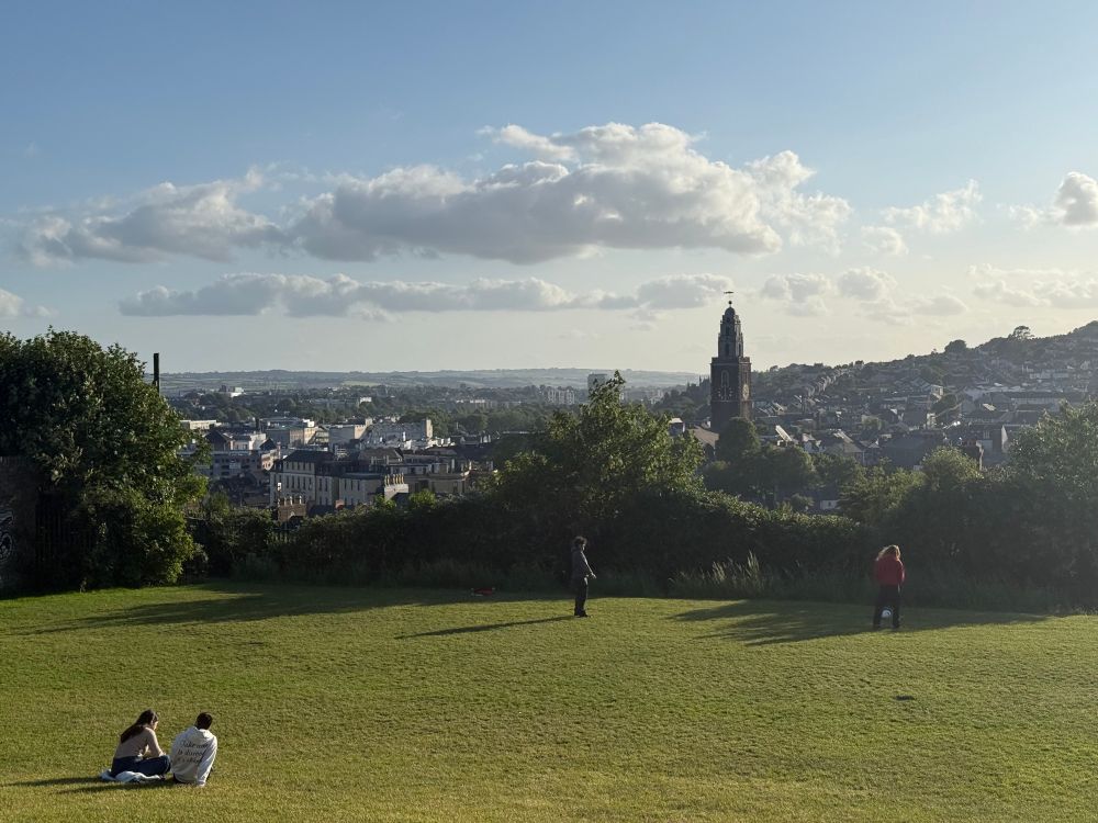 Panoramic view of Cork, Ireland, on a sunny day with a blue sky and scattered clouds. In the foreground, two people are sitting on the grass to the left, while two others stand further back on the lawn. In the background, the cityscape of Cork is visible, featuring the prominent tower of St. Anne’s Church (Shandon) at the center, surrounded by houses and buildings, with green hills rising in the distance.